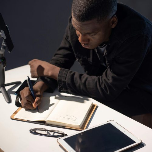 A man in concentration writing notes at a desk with a tablet nearby, indoors setting.